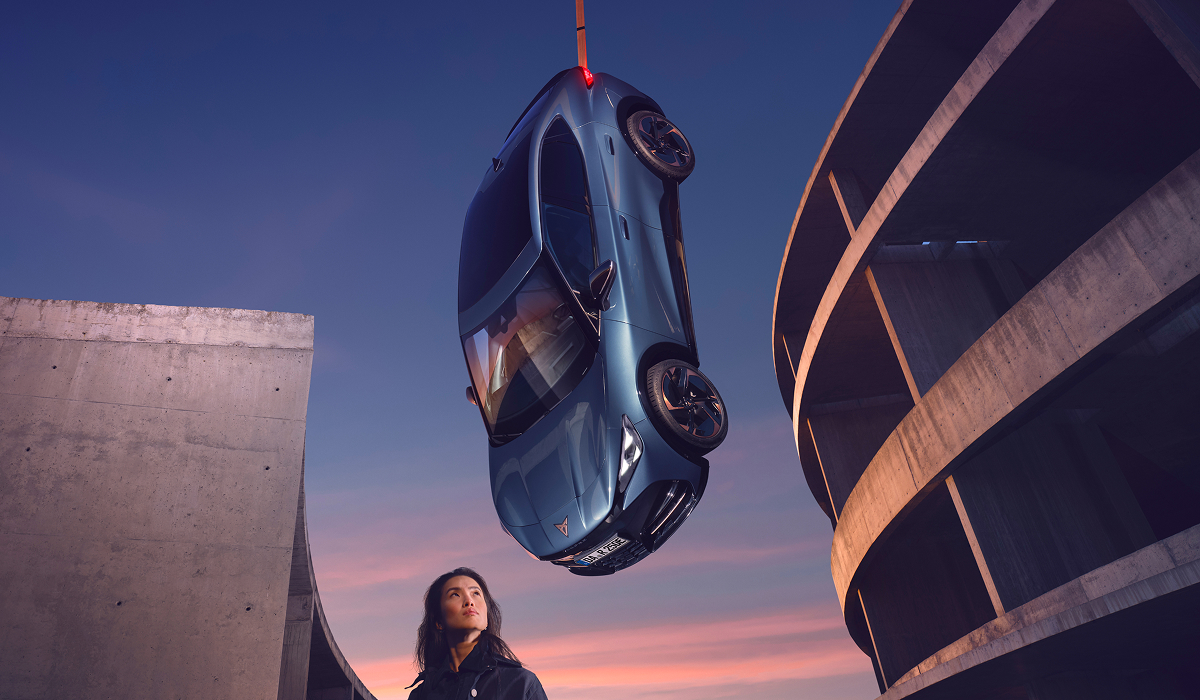 Frau blickt nach oben auf ein hängenden CUPRA Tavascan, das an einem Kran vor einer Betonarchitektur schwebt